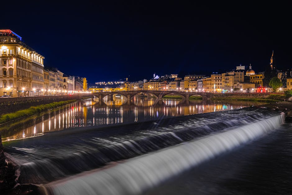 A nighttime view of a cityscape with illuminated historic buildings along a river, reflecting their warm lights on the water's surface. The river features a small weir with smooth, flowing water casca